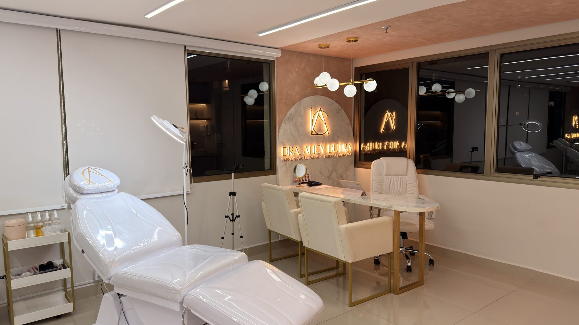 Sala de atendimento com maca branca, mesa de mármore e painel iluminado com o nome da Dra. Alicy Dutra